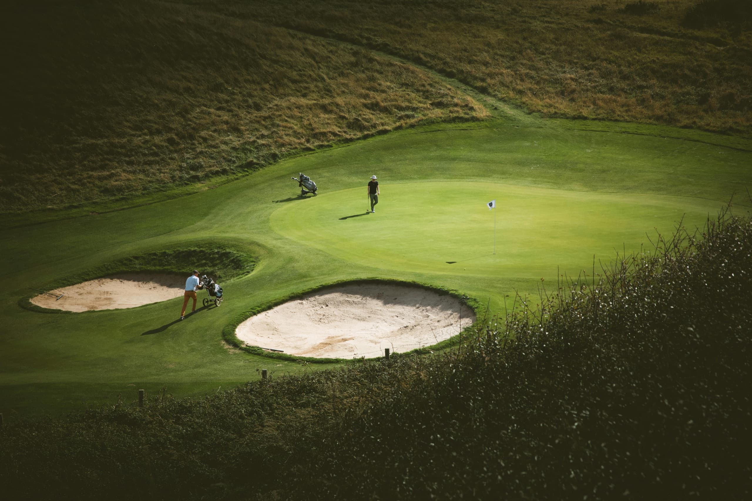 An elevated, sunlit view of a college course green — two players walking the putting surface beside bunkers, flag in place